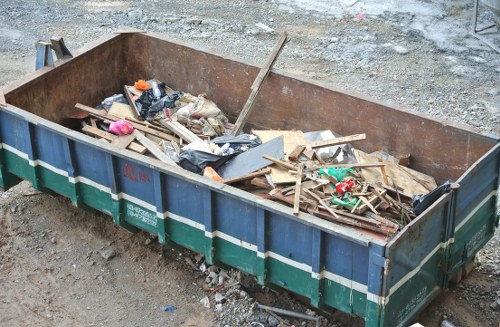 A large rectangular metal skip with a rusted exterior and green and white striped sides is positioned on an uneven gravel surface, filled with various types of construction and garden waste including wooden planks, broken bricks, plastic bags, and miscellaneous debris. The skip's interior contains a mixture of raw timber, some cut and weathered, alongside black plastic rubbish bags that are partially torn open, revealing more waste materials inside. Surrounding the skip, the ground appears rough with loose gravel and dirt, indicative of a construction or garden clearance site in West Ham. The setting is outdoors, with no other objects or structures visible, emphasizing the focus on the skip's contents. Garden Clearance West Ham's service description encompasses rubbish removal, including debris like this typically generated in garden or property clearance projects, highlighting their capacity to manage bulky waste efficiently in the local area.