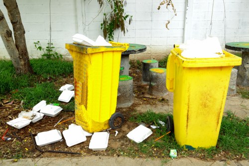 Two large yellow wheelie bins, positioned on a sidewalk in an outdoor urban area, with their lids open and overflowing with white foam food containers. Several more foam containers are scattered on the ground around the bins, some lying on their sides and others partially crushed. The scene is set against a white wooden fence with visible horizontal panels, and there are small patches of grass and weeds near the base of a tree trunk on the left. In the background, behind the bins, there are short concrete stools with painted tops in yellow and green, arranged on a paved surface. The environment appears to be a residential or community area in West Ham, indicative of typical waste disposal issues, with a focus on rubbish related to food packaging. The overall scene reflects improper waste disposal, highlighting the services provided by Garden Clearance West Ham for rubbish removal and garden clearance tasks.