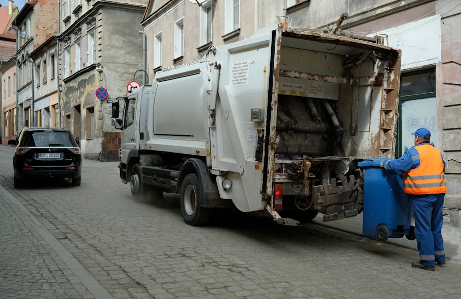 A pile of mixed rubbish, including cardboard boxes, paper bags, and plastic packaging, is gathered on the ground next to a brick wall and beneath a large tree trunk in an outdoor setting. The boxes ar