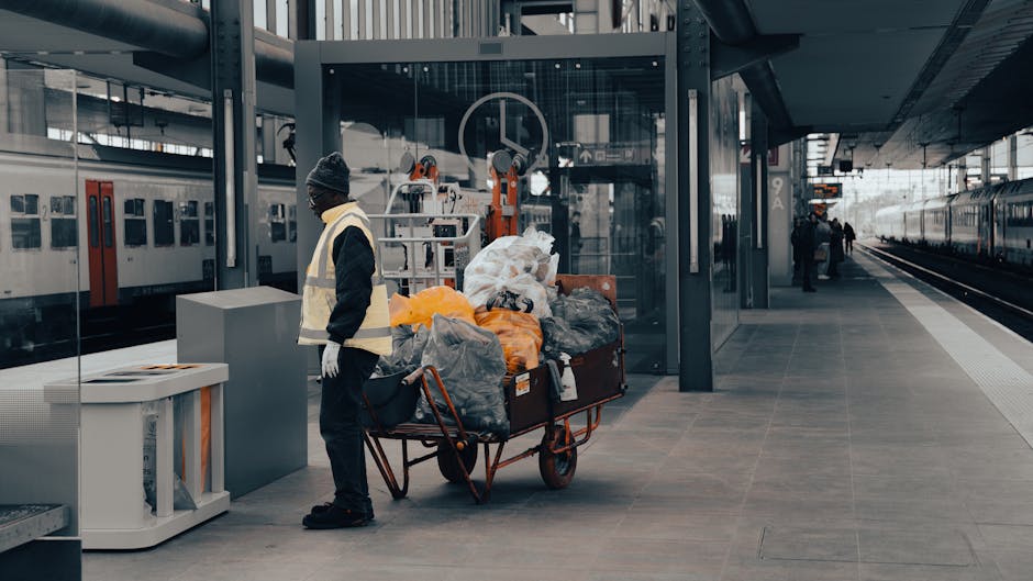 An urban street scene showing a waste collection truck operated by a waste management worker in a high-visibility orange and blue uniform, standing beside the vehicle. The truck, which is white with v