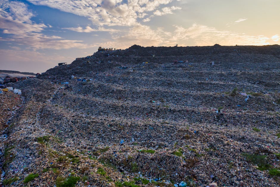 An expansive aerial view of a large landfill site filled with mixed household and commercial waste materials, including plastic packaging, paper, textiles, and organic matter. The waste is piled dense